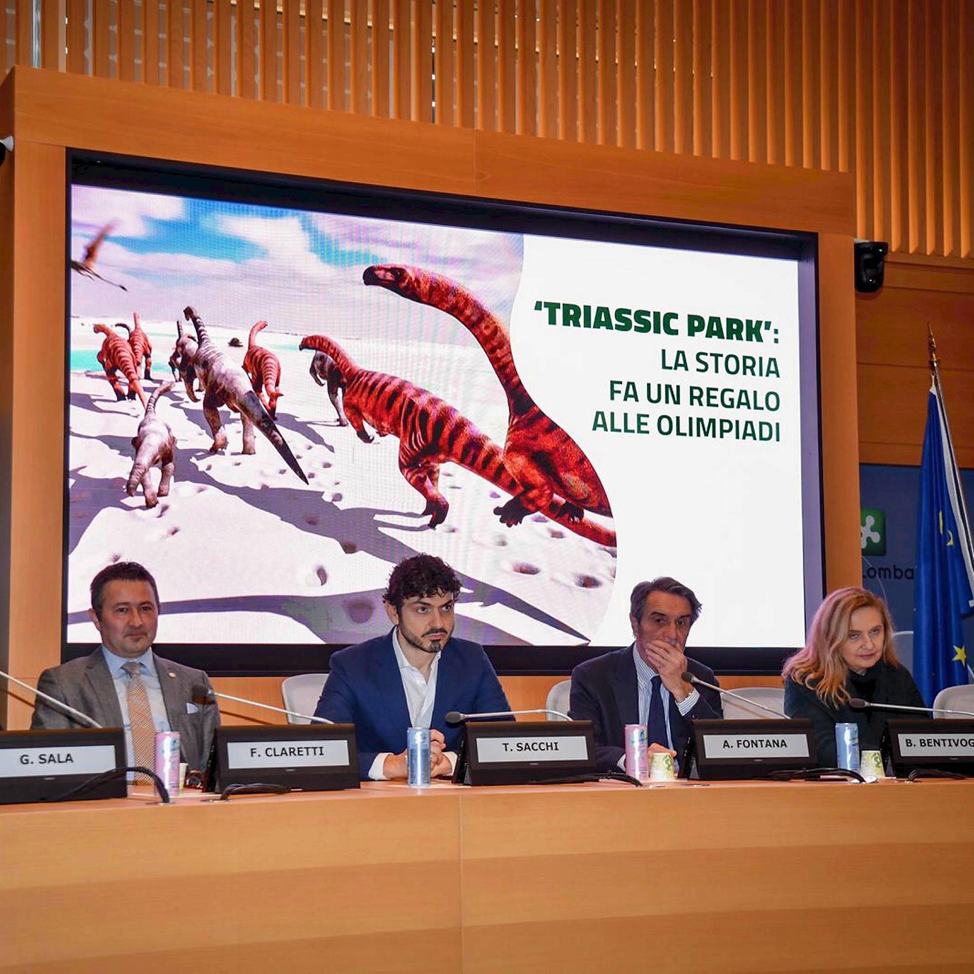 Mehrere Menschen sitzen bei einer Pressekonferenz an einem Tisch mit Mikrofonen. Im Hintergrund ist eine Leinwand mit einer computergenerierten Grafik von Dinosauriern und dem Titel "Triassic Park: Die Geschichte beschenkt die Olympischen Spiele".