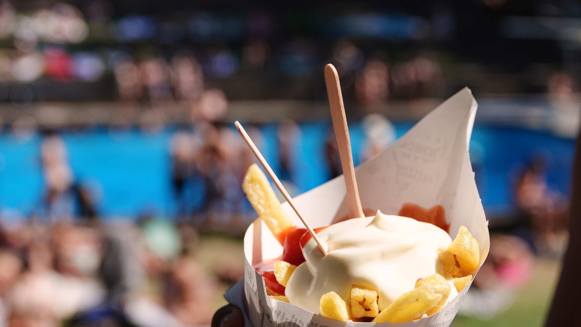 Eine Tüte Pommes Frites, im Hintergrund Menschen in einem Freibad.