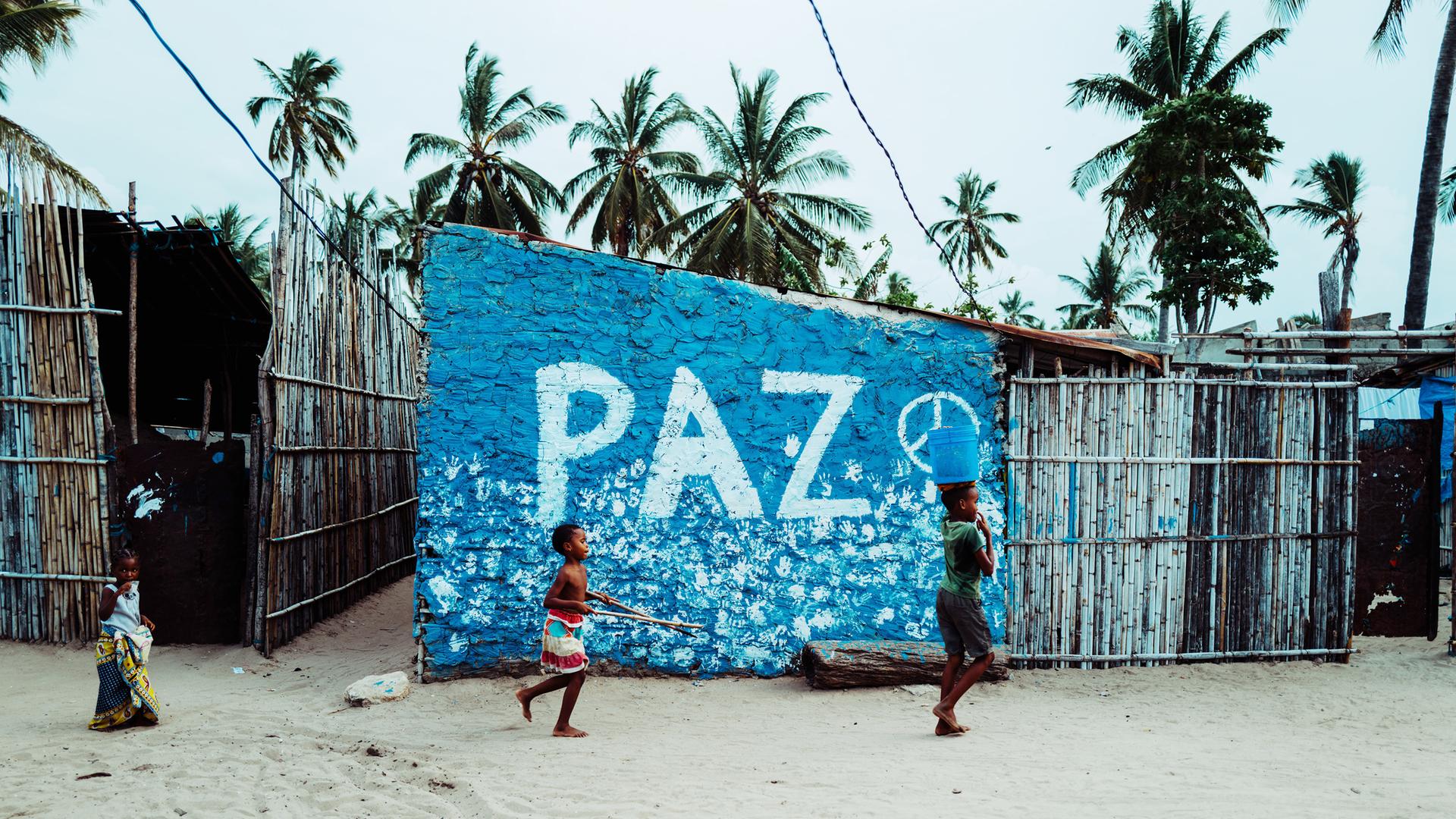 Eine Blaue Wand mit dem Wort "PAZ" (Frieden) in Cabo Delgado, Mosambik. Kinder spielen davor.