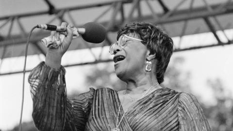 Eine Sängerin steht auf einer Bühne und gibt ein Konzert. Es ist die Jazz-Musikerin Ella Fitzgerald. Sie hält ein Mikrofon in der Hand. Es ist eine Schwarz-weiß-Aufnahme.