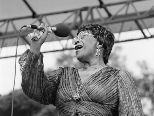 Eine Sängerin steht auf einer Bühne und gibt ein Konzert. Es ist die Jazz-Musikerin Ella Fitzgerald. Sie hält ein Mikrofon in der Hand. Es ist eine Schwarz-weiß-Aufnahme.