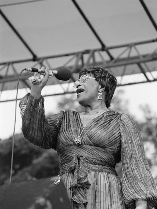 Eine Sängerin steht auf einer Bühne und gibt ein Konzert. Es ist die Jazz-Musikerin Ella Fitzgerald. Sie hält ein Mikrofon in der Hand. Es ist eine Schwarz-weiß-Aufnahme.