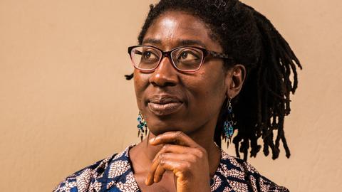 Gloria Boateng stützt das Kinn auf ihre Hand. Sie trägt eine blau gemusterte Bluse und das schwarze Haar in Braids.