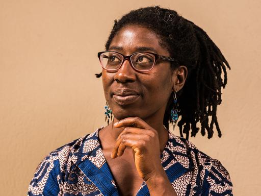 Gloria Boateng stützt das Kinn auf ihre Hand. Sie trägt eine blau gemusterte Bluse und das schwarze Haar in Braids.