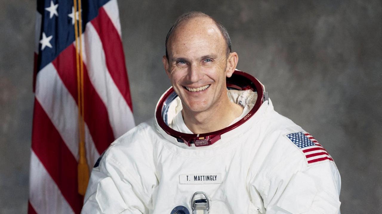 Der Astronaut Thomas K. Mattingly steht in einem Raumanzug vor einer US-amerikanischen Fahne und lächelt in die Kamera.