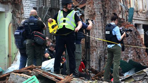 Ein Polizist sichert die Arbeit von Reportern ab, die das zerstörte Kulturhaus in Tschuhujiw fotografieren. Es war von einer russischen Rakete getroffen worden. Die Fotografen richten ihre Apparate auf die Trümmer, der Polizist steht im Vordergrund und trägt eine gelbe Warnweste.