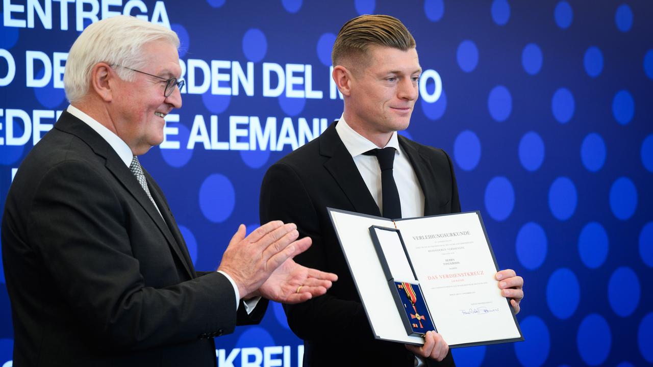 Die beiden stehen vor einer dunkelblauen Wand mit hellblauen Kreisen und weißer Schrift. Sie tragen beide schwarze Jacketts. Kroos präsentiert die offene Schatulle mit dem Verdienstkreuz und die dazugehörige Urkunde. 