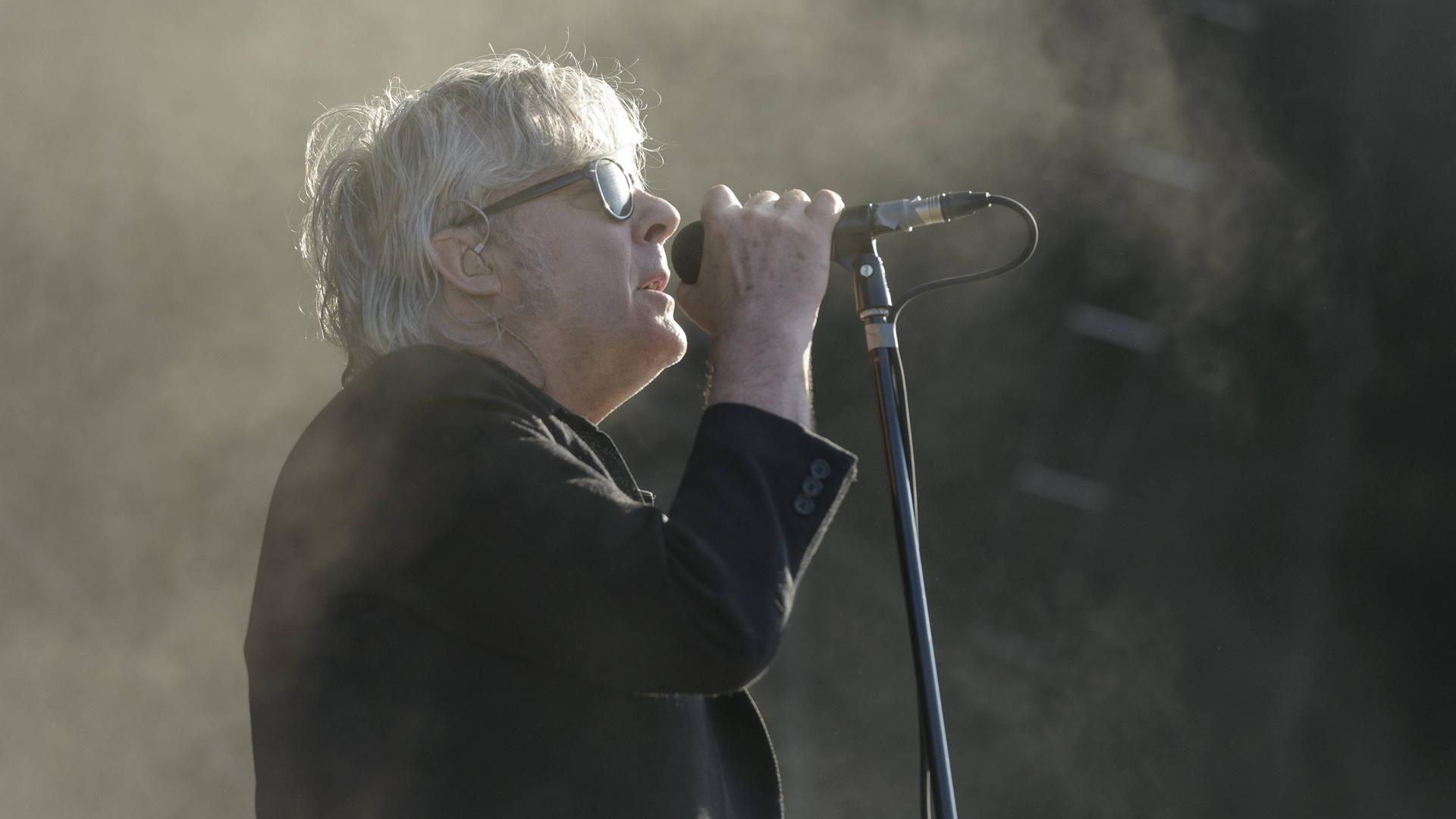 Ein Mann mit weiß-grauen Haaren, Sonnenbrille und schwarzem Jacket steht im rechten Seitenprofil mit der rechten Hand am Mikrofon und singt.