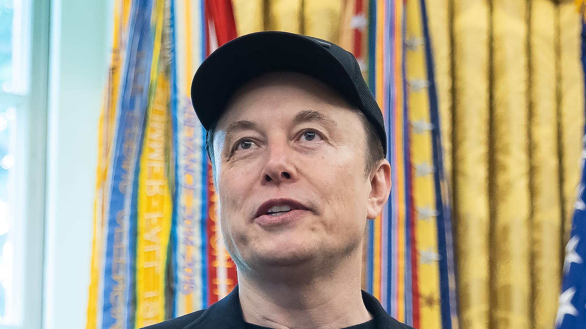 Das Bild zeigt ein Porträt von Elon Musk. Er lächelt und trägt eine dunkle Baseballkappe.