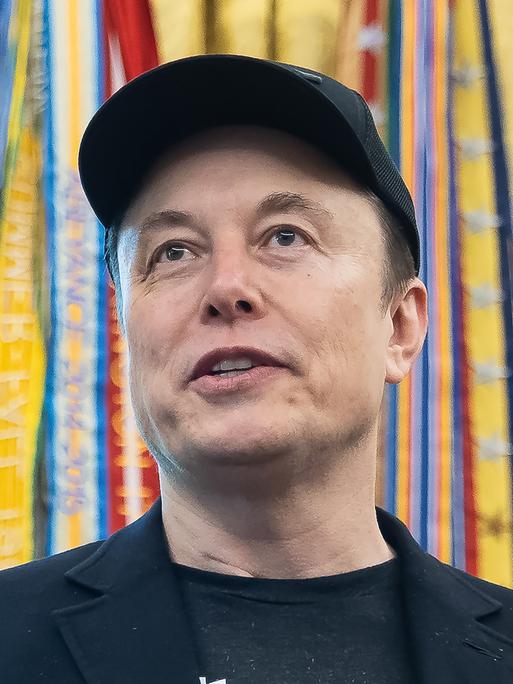 Das Bild zeigt ein Porträt von Elon Musk. Er lächelt und trägt eine dunkle Baseballkappe.