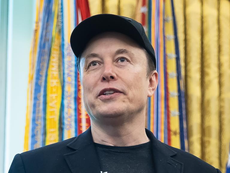 Das Bild zeigt ein Porträt von Elon Musk. Er lächelt und trägt eine dunkle Baseballkappe.