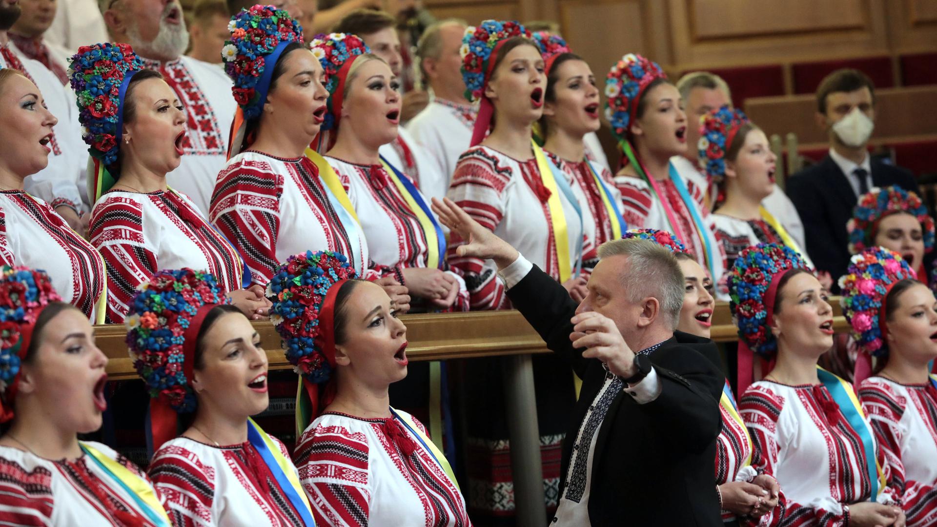 Der Dirigent Zenovii Korinets dirigiert einen Chor, deren Mitglieder traditionelle Trachten tragen. Die Frauen haben dabei bunte Kronen auf dem Haupt.