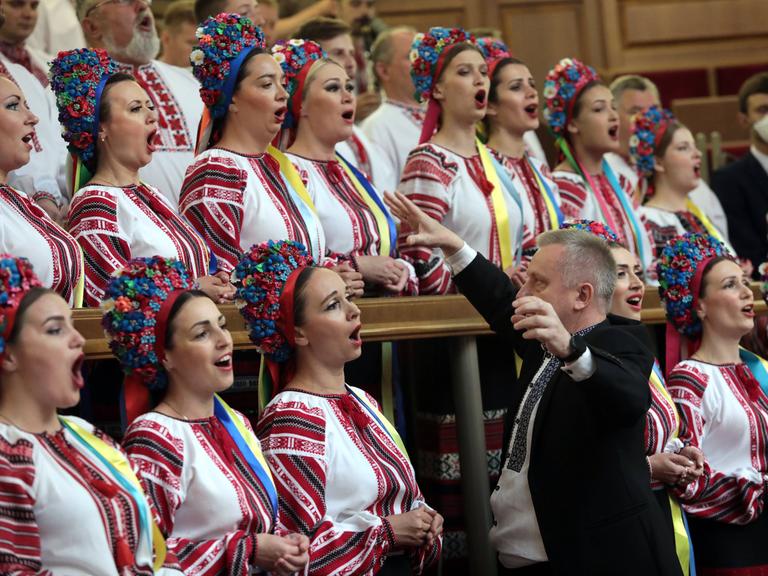 Der Dirigent Zenovii Korinets dirigiert einen Chor, deren Mitglieder traditionelle Trachten tragen. Die Frauen haben dabei bunte Kronen auf dem Haupt.