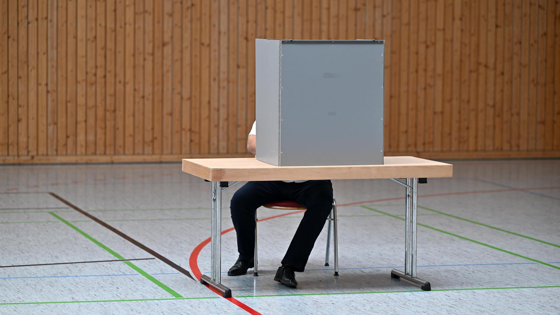 Ein Mann sitzt verdeckt hinter einer Wahlkabine in einem Wahllokal in einer Turnhalle. 