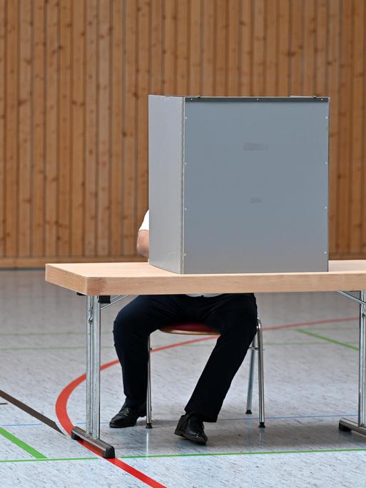 Ein Mann sitzt verdeckt hinter einer Wahlkabine in einem Wahllokal in einer Turnhalle. 