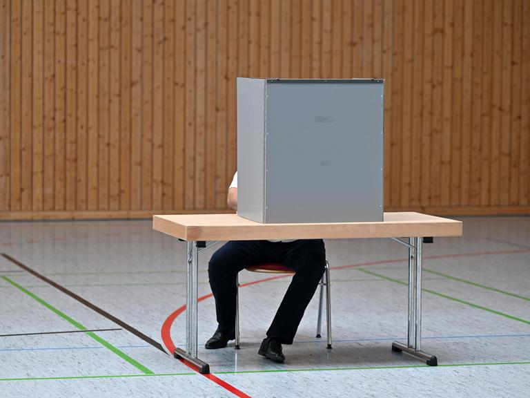 Ein Mann sitzt verdeckt hinter einer Wahlkabine in einem Wahllokal in einer Turnhalle. 