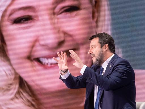 Ein Mann mit blauem Anzug sitzt vor einem großen Bildschirm und gestikuliert. Es ist der italienische Politiker Matteo Salvini. Im Hintergrund auf dem Bildschirm ist eine blonde Frau, die französische Politikerin Marine Le Pen.