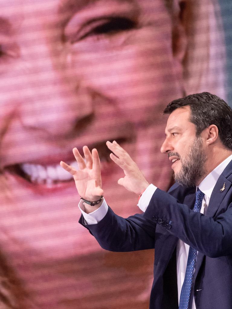 Ein Mann mit blauem Anzug sitzt vor einem großen Bildschirm und gestikuliert. Es ist der italienische Politiker Matteo Salvini. Im Hintergrund auf dem Bildschirm ist eine blonde Frau, die französische Politikerin Marine Le Pen.