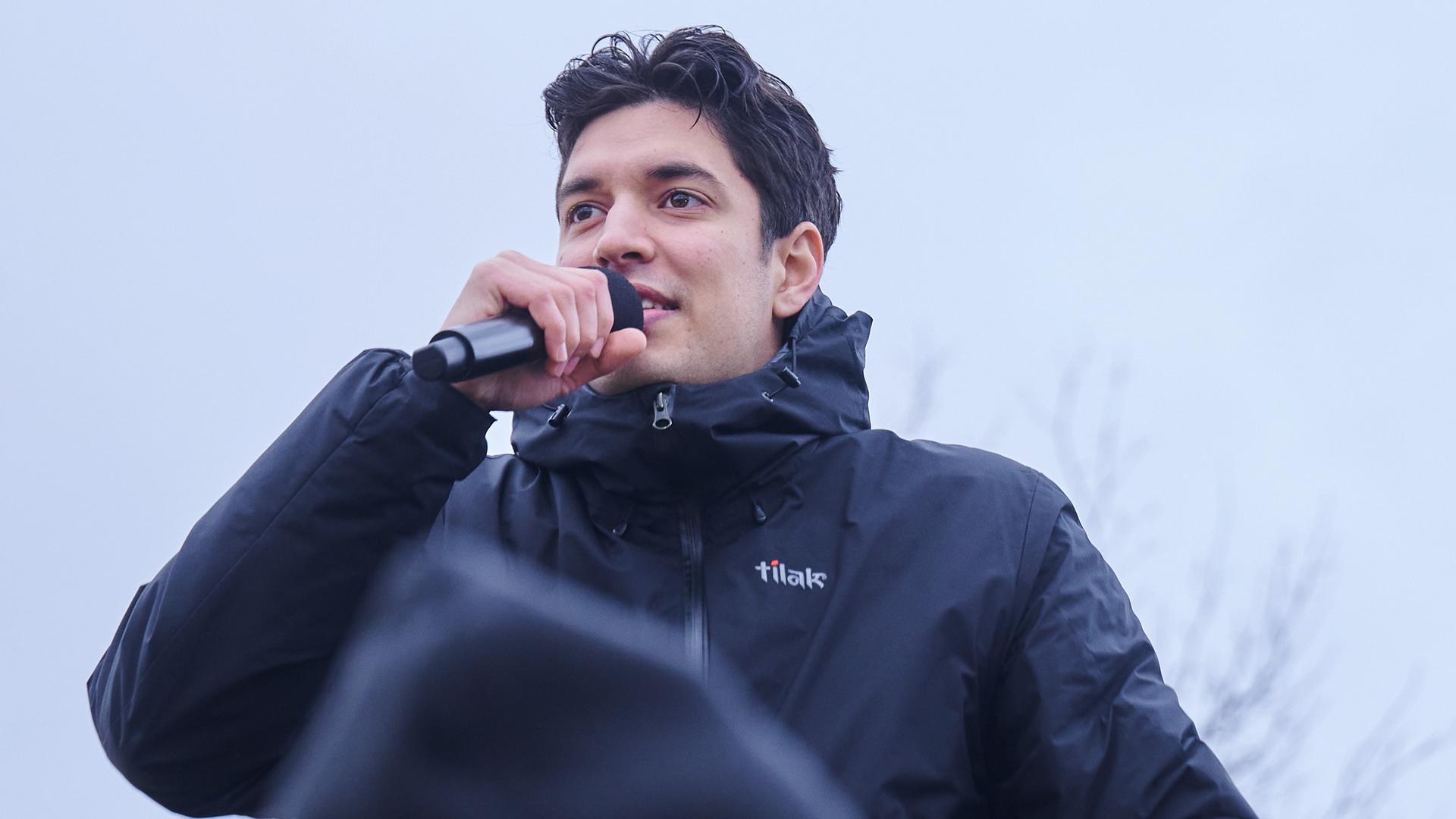 Apsilon, Influencer und Sänger singt während einer Demonstration gegen rechts auf einer Bühne. Er trägt eine schwarze Jacke und hält ein Handmikrofon vor den Mund. 