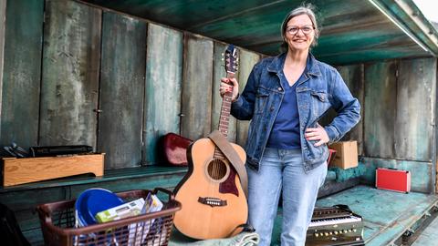 Brigitte Dethier hält eine Gitarre in der Hand und lacht in die Kamera. Um sie herum liegen verschiedene Theaterrequisiten. 