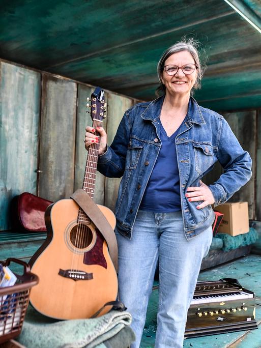 Brigitte Dethier hält eine Gitarre in der Hand und lacht in die Kamera. Um sie herum liegen verschiedene Theaterrequisiten. 
