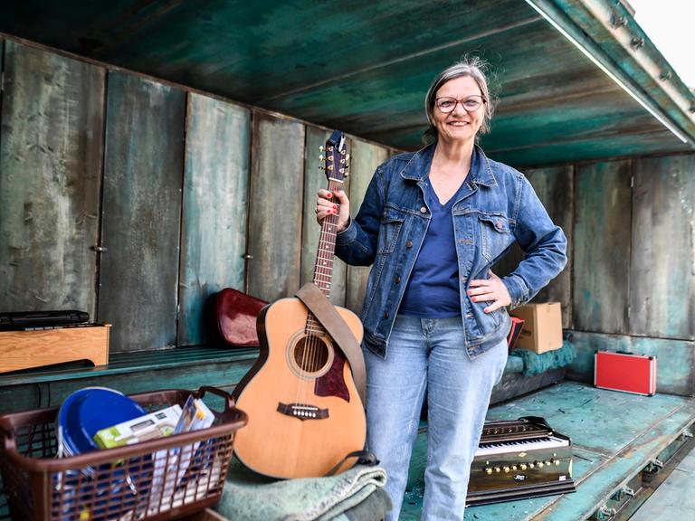 Brigitte Dethier hält eine Gitarre in der Hand und lacht in die Kamera. Um sie herum liegen verschiedene Theaterrequisiten. 