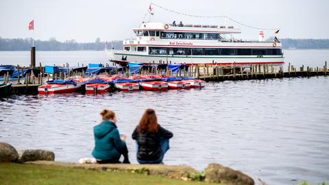 Zwei Frauen sitzen am Ufer, während das Ausflugsschiff bei sonnigem Wetter zu Beginn einer Rundfahrt über das Zwischenahner Meer von einem Steg ablegt.