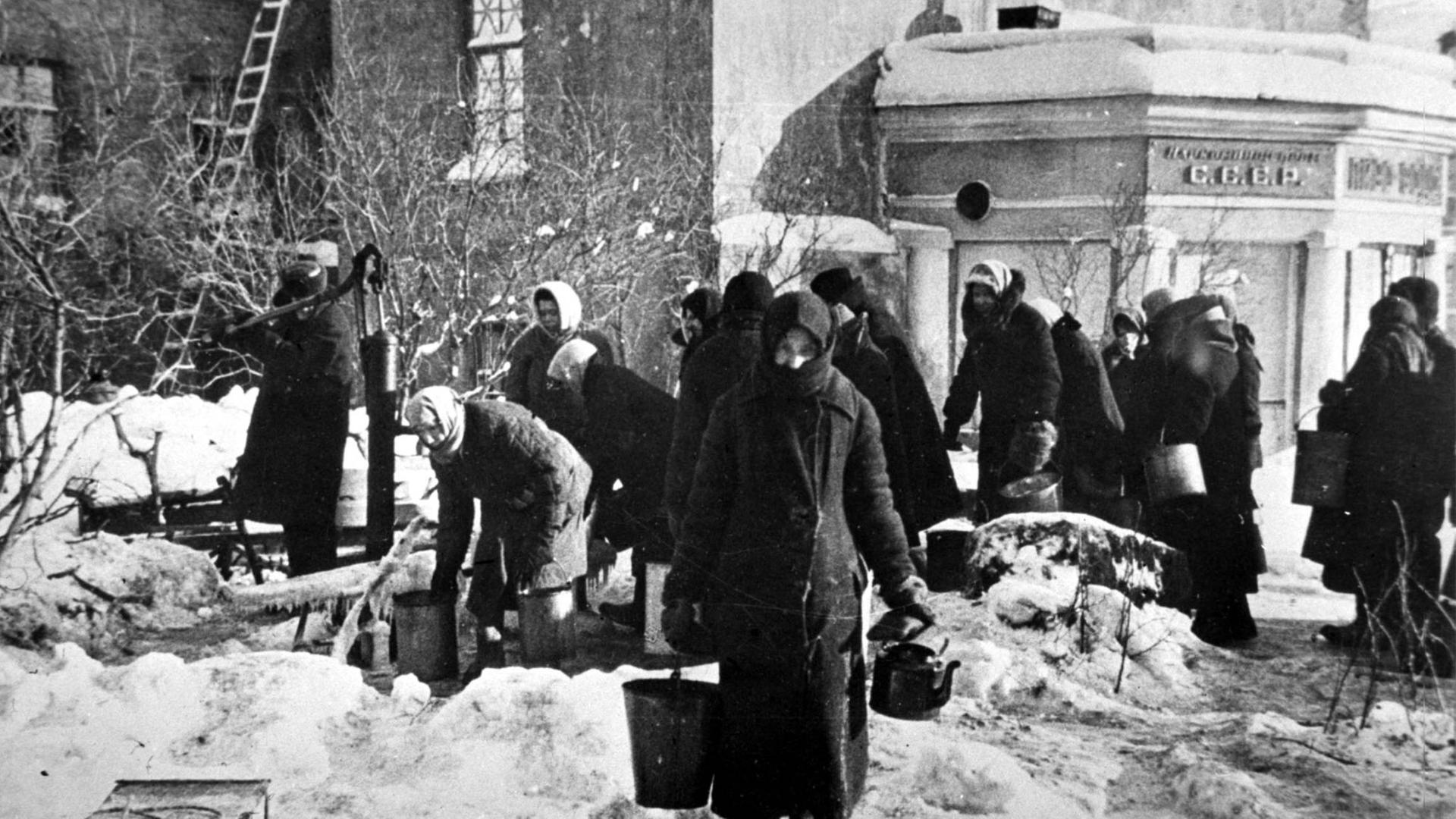 Schwarz-weiß-Aufnahme aus Leningrad im Winter 1942/1943. Mehrere Frauen versammeln sich an einer Wasserpumpe, im Hintergrund ein Haus.