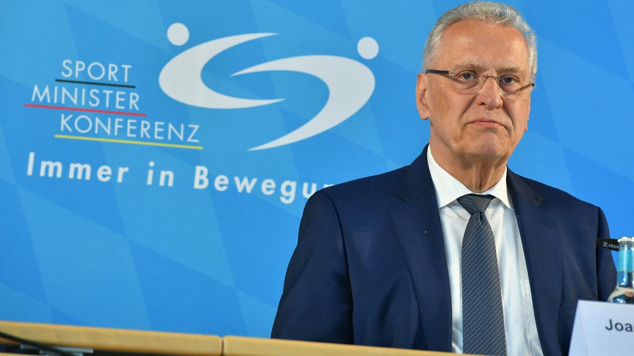 Bayerns Sportminister Herrmann sitzt an einem Tisch, im Hintergrund ist eine blaue Wand zu sehen, auf der untereinander geschrieben die Wörter stehen: Sport, Minister und Konferenz.