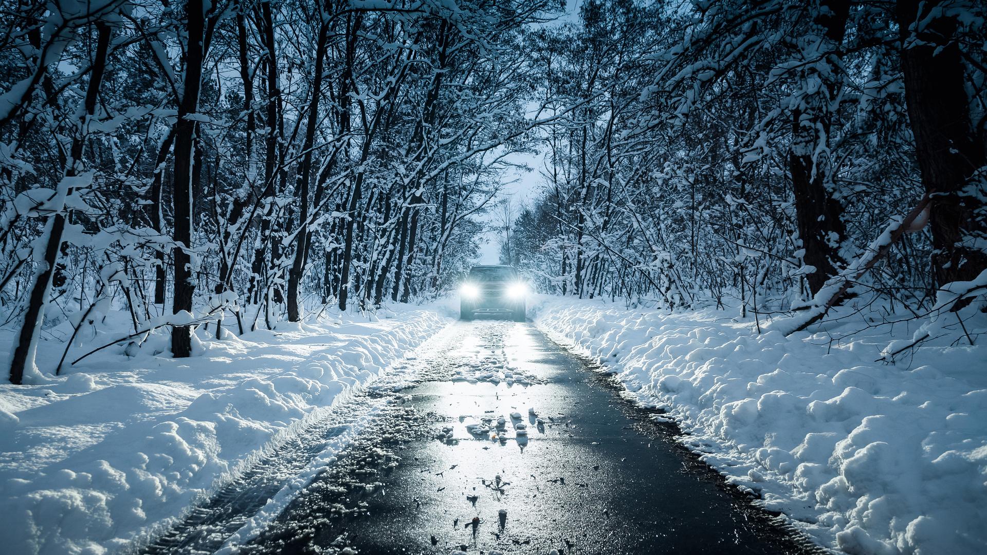 Scheinwerfer von einem Auto auf einer schmalen schneebedeckten Straße durch einen Wald.