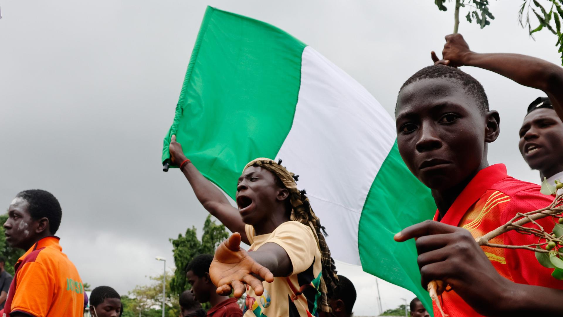 Ein Jugendlicher hält die grün-weiße nigerianische Flagge in der Hand und schreit. Rechts neben ihm blickt ein weiterer Jugendlicher in die Kamera.