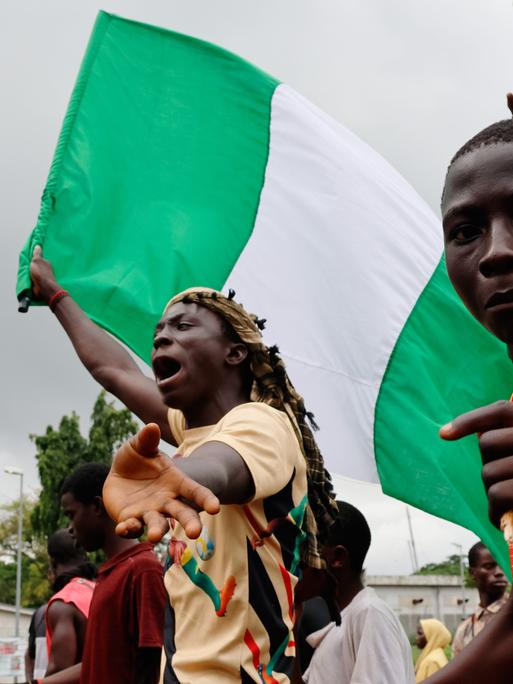 Ein Jugendlicher hält die grün-weiße nigerianische Flagge in der Hand und schreit. Rechts neben ihm blickt ein weiterer Jugendlicher in die Kamera.