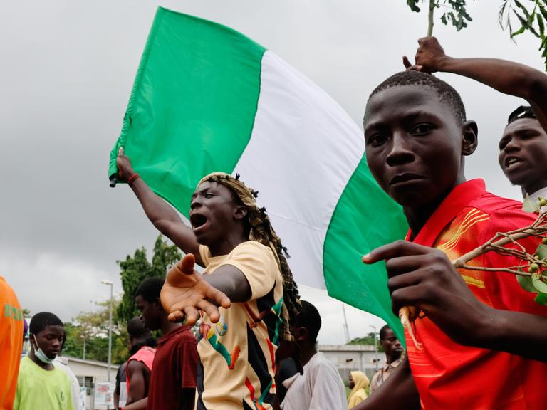 Ein Jugendlicher hält die grün-weiße nigerianische Flagge in der Hand und schreit. Rechts neben ihm blickt ein weiterer Jugendlicher in die Kamera.