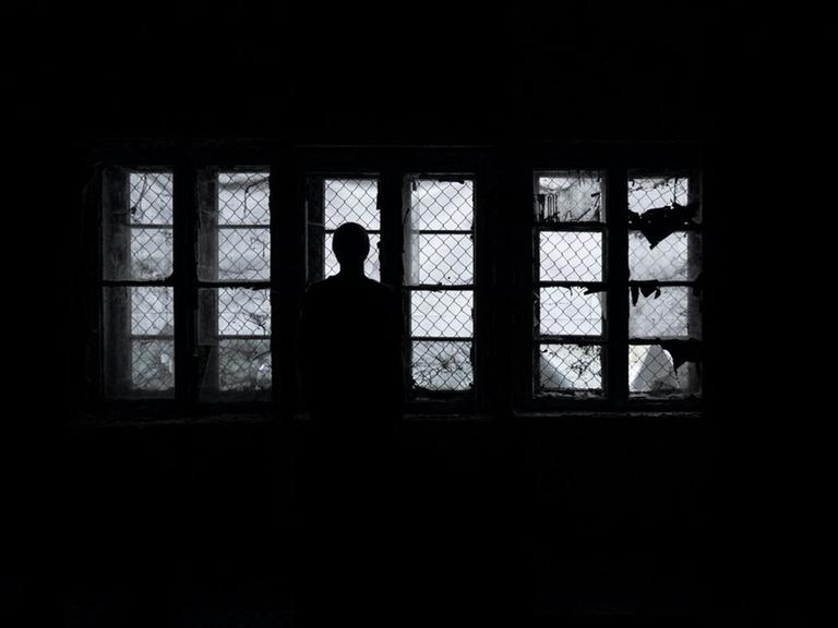 Eine Frau steht als Silhouette im Dunkeln vor vergitterten Fenstern und schaut hinaus