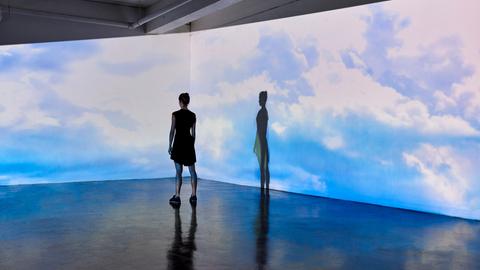 Eine junge Frau in einem Museum schaut auf eine Wand, an die große Bilder vom bewölkten Himmel projiziert werden.