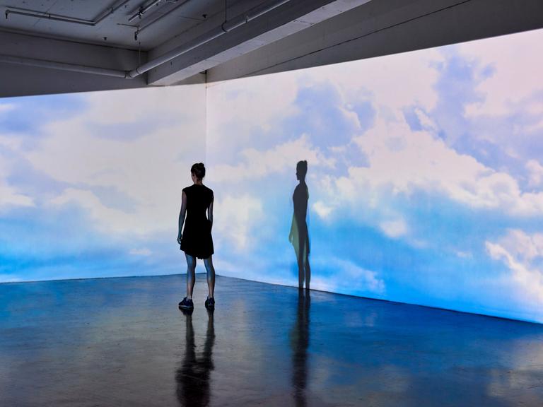 Eine junge Frau in einem Museum schaut auf eine Wand, an die große Bilder vom bewölkten Himmel projiziert werden.