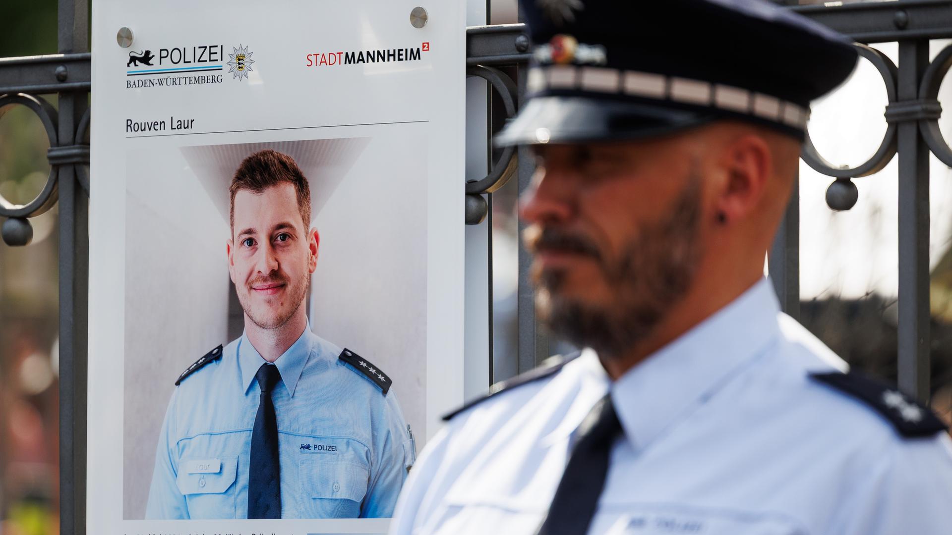 Ein Polizist steht neben einer Gedenktafel für Rouven Laur in Mannheim. Auf der Tafel ist ein Bild des getöteten Polizisten. Ein Polizist steht neben einer Gedenktafel für Rouven Laur in Mannheim. Auf der Tafel ist ein Bild des getöteten Polizisten.
