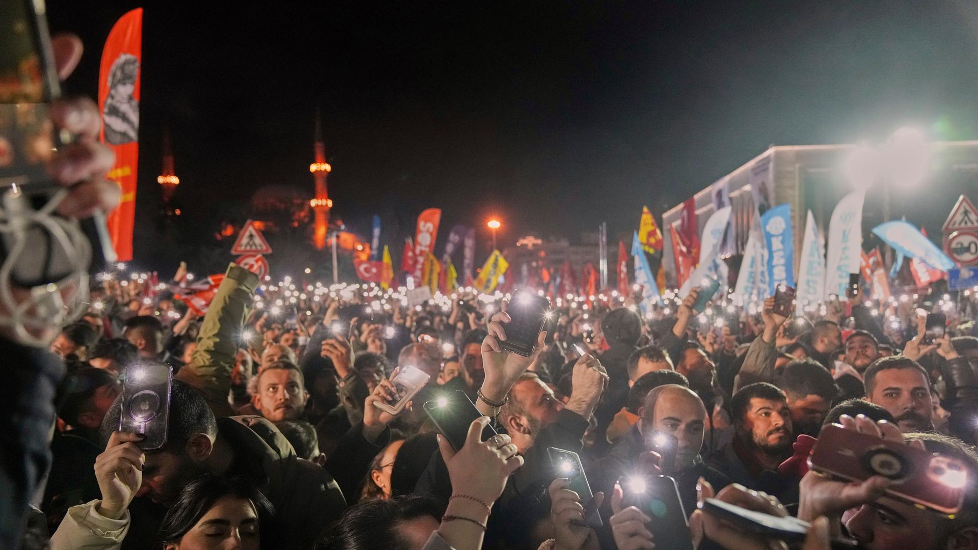 Eine Menschenmenge steht im Dunklen vor der Stadtverwaltung in Istanbul. Die Protestteilnehmenden recken ihre Smartphones mit angeschalteter Taschenlampenfunktion in die Höhe. Im Hintergrund sind Fahnenaufsteller zu sehen. 