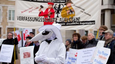 Eine Person mit Maske steht vor einer Menschenmenge mit Protestschildern.