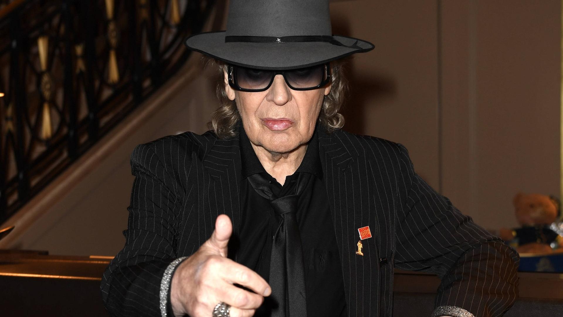Udo Lindenberg lehnt schwarz gekleidet sowie mit schwarzem Hut und dunkler Brille an einem Tresen und blickt zufrieden in die Kamera.