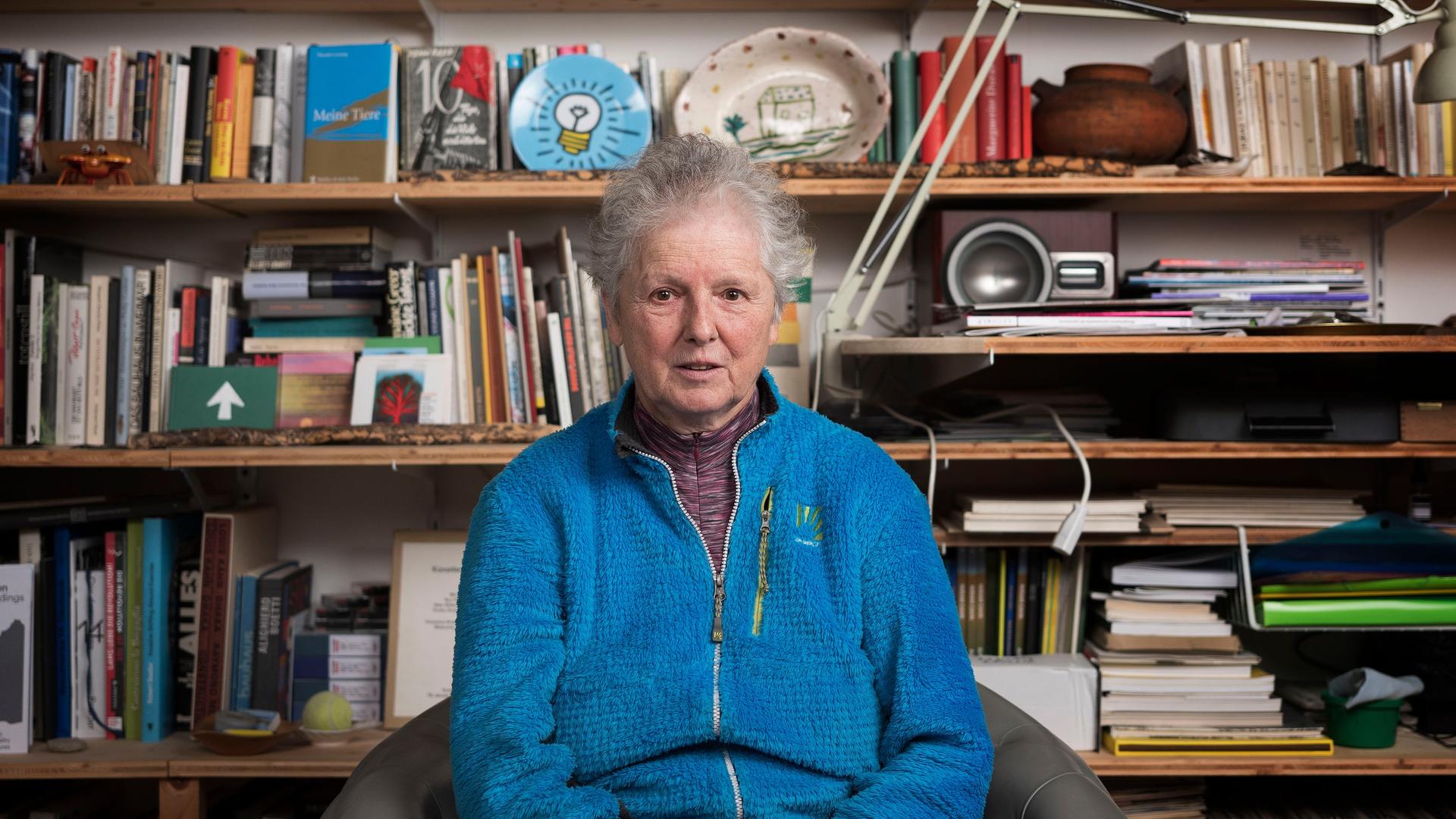 Miriam Cahn sitzt vor ihrem Bücherregal. Sie trägt kurzes graues Haar und eine leuchtend blaue Jacke.