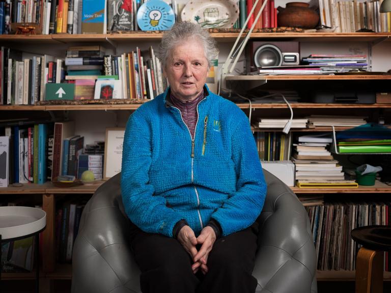 Miriam Cahn sitzt vor ihrem Bücherregal. Sie trägt kurzes graues Haar und eine leuchtend blaue Jacke.