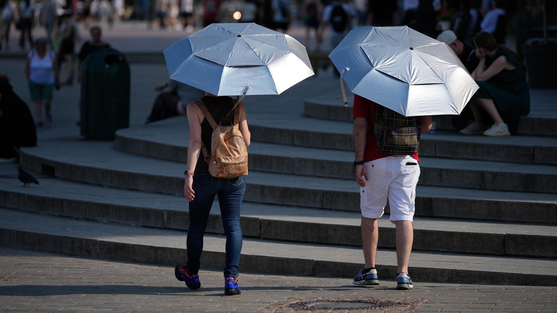 Zwei Menschen laufen mit silbernen UV-Sonnenschirmen durch eine Innenstadt während der Hitzewelle im Sommer 2026.