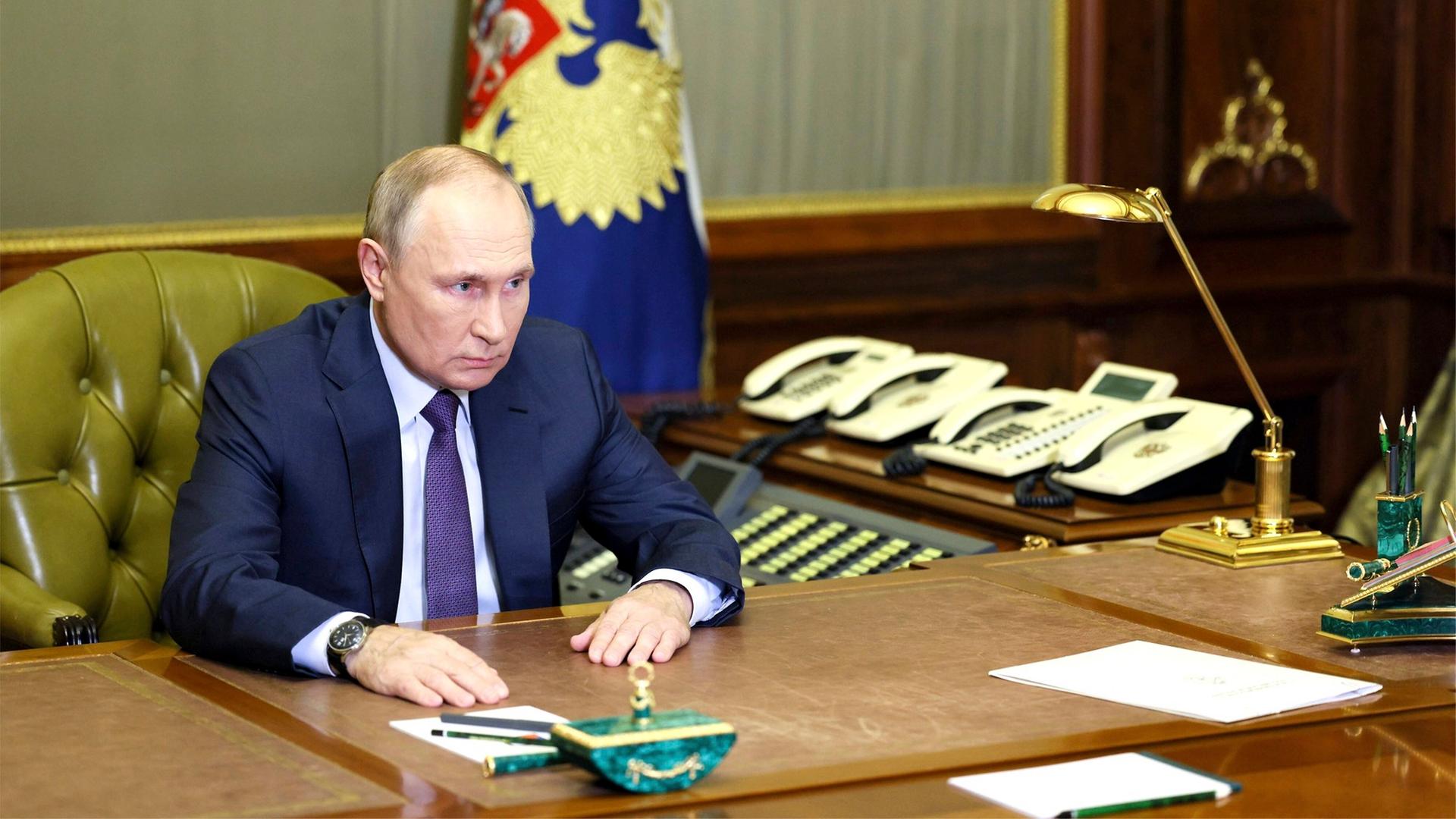 Der russische Präsident Putin, allein an seinem Schreibtisch. Im Hintergrund stehen eine Handvoll Telefone nebeneinander aufgereiht.