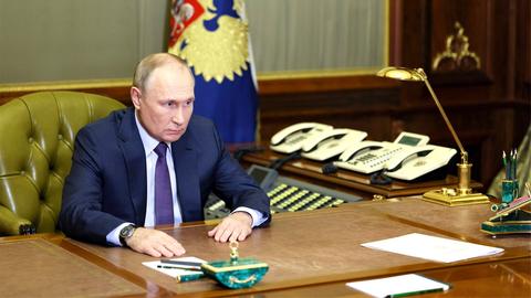 Der russische Präsident Putin, allein an seinem Schreibtisch. Im Hintergrund stehen eine Handvoll Telefone nebeneinander aufgereiht.