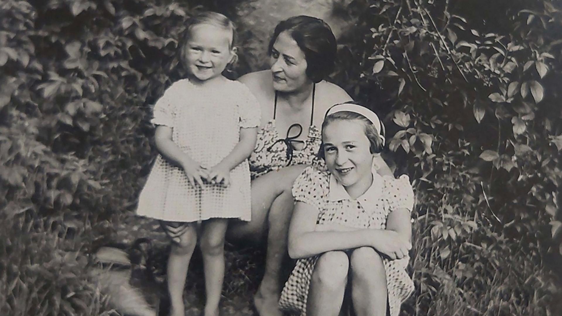 Zwischen zwei großen Büschen sitzt Eva mit ihrer Schwester Yvonne und ihrer Mutter. Die beiden Mädchen lachen den Fotografen an, die Mutter schaut liebevoll auf Eva. Zwischen zwei großen Büschen sitzt Eva mit ihrer Schwester Yvonne und ihrer Mutter. Die beiden Mädchen lachen den Fotografen an, die Mutter schaut liebevoll auf Eva.