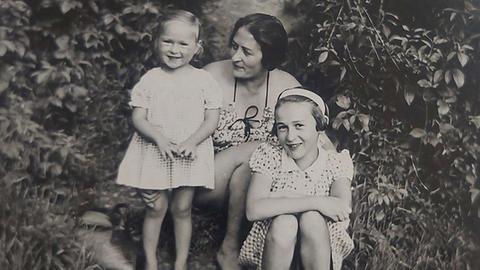 Zwischen zwei großen Büschen sitzt Eva mit ihrer Schwester Yvonne und ihrer Mutter. Die beiden Mädchen lachen den Fotografen an, die Mutter schaut liebevoll auf Eva. Zwischen zwei großen Büschen sitzt Eva mit ihrer Schwester Yvonne und ihrer Mutter. Die beiden Mädchen lachen den Fotografen an, die Mutter schaut liebevoll auf Eva.