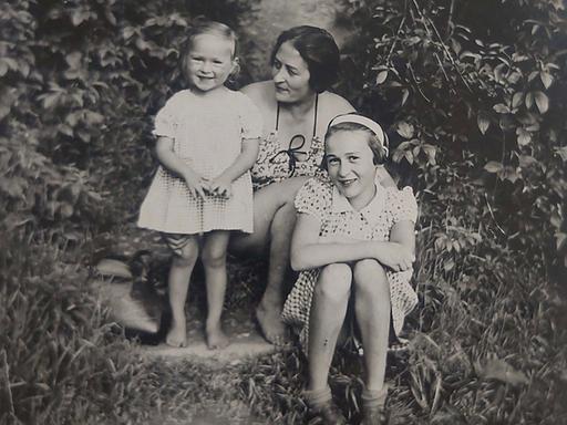 Zwischen zwei großen Büschen sitzt Eva mit ihrer Schwester Yvonne und ihrer Mutter. Die beiden Mädchen lachen den Fotografen an, die Mutter schaut liebevoll auf Eva.