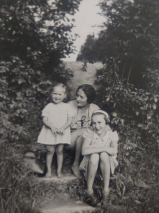 Zwischen zwei großen Büschen sitzt Eva mit ihrer Schwester Yvonne und ihrer Mutter. Die beiden Mädchen lachen den Fotografen an, die Mutter schaut liebevoll auf Eva.