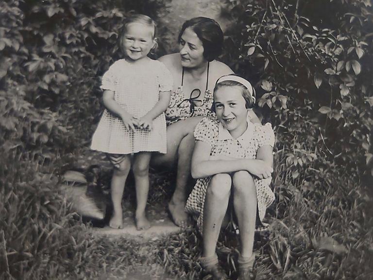 Zwischen zwei großen Büschen sitzt Eva mit ihrer Schwester Yvonne und ihrer Mutter. Die beiden Mädchen lachen den Fotografen an, die Mutter schaut liebevoll auf Eva.